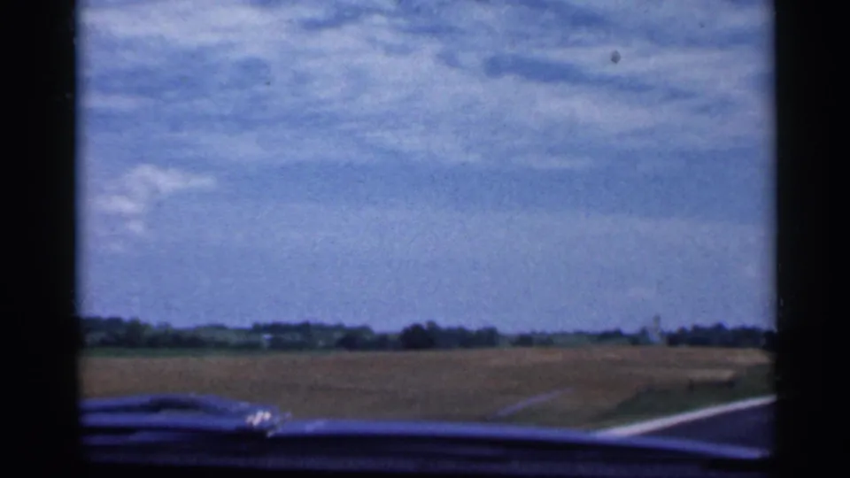 1959 Indiana home-movie frame showing a passenger view of a rural road, open fields, and scattered clouds through the car windshield.
