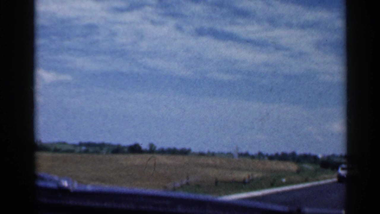 1959 Indiana driving view with a two-lane road, fields, distant trees, and a car partly visible ahead on the right.