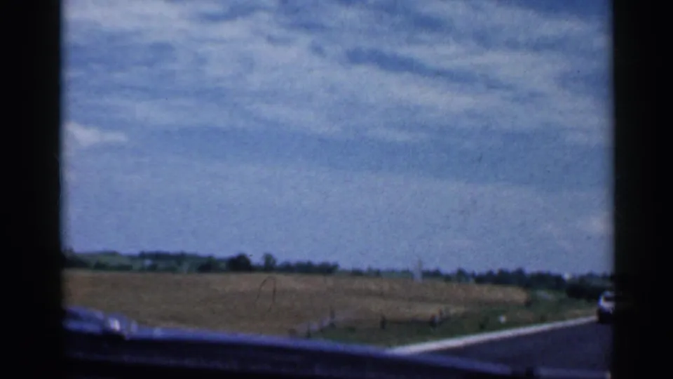 1959 Indiana driving view with a two-lane road, fields, distant trees, and a car partly visible ahead on the right.
