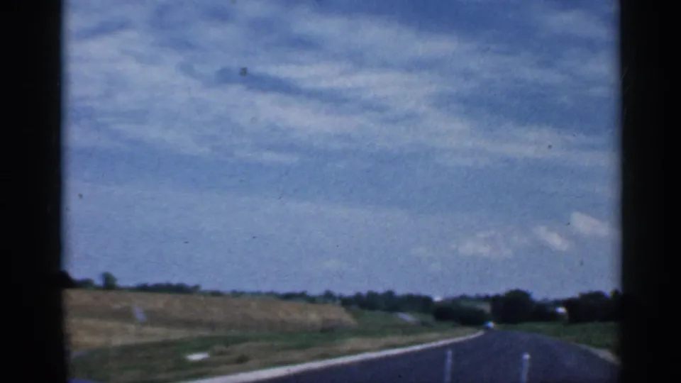 Passenger-view frame from a 1959 Indiana rural drive, showing a paved road, fields, and a broad sky from inside the moving car.