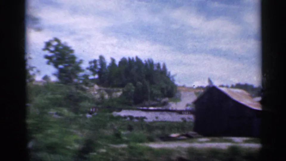 Indiana rural scene with a dark outbuilding, water or wet ground, trees, and cloudy sky in a 1959 home movie frame.