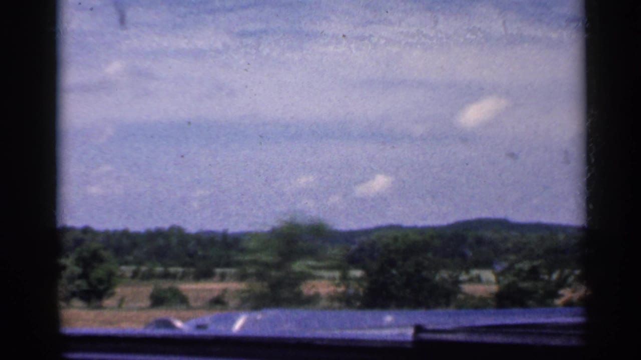 1959 Indiana car-window view with the dashboard edge visible below distant fields, wooded hills, and scattered clouds.