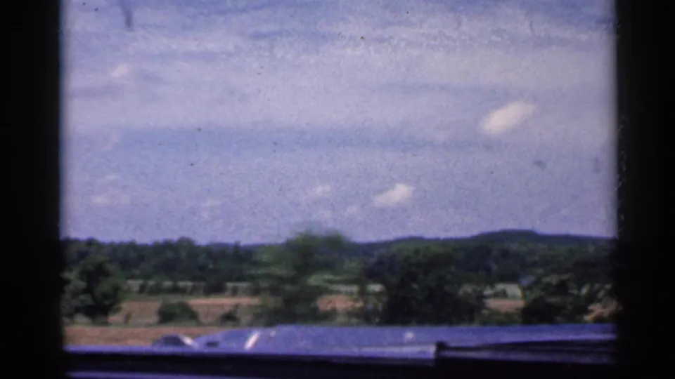 1959 Indiana car-window view with the dashboard edge visible below distant fields, wooded hills, and scattered clouds.