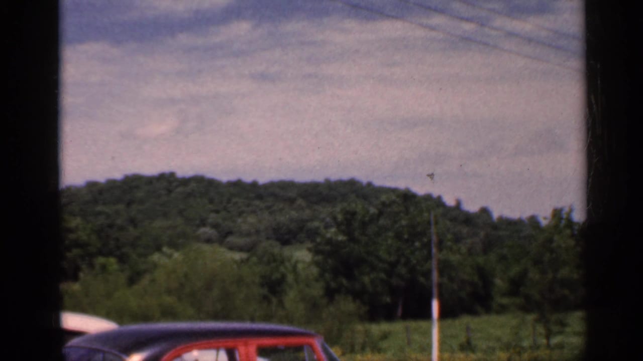 1959 Indiana roadside view with a wooded hill, open field, utility pole, and the top of a red mid-century car in frame.