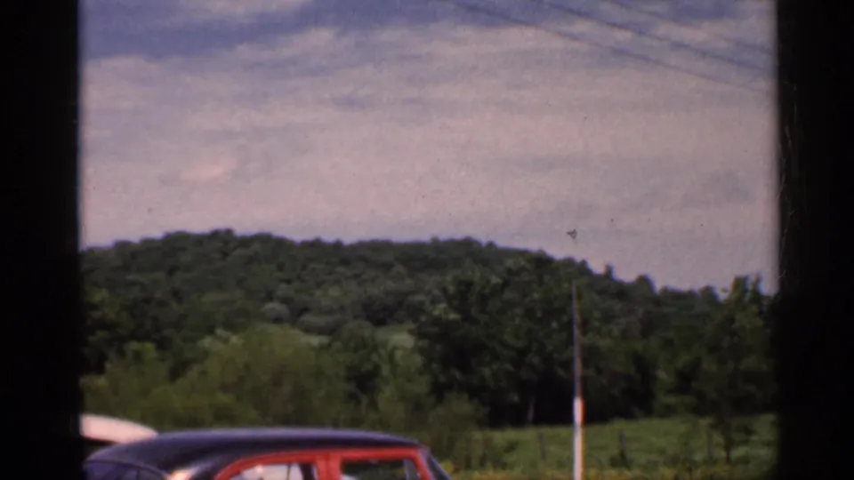 1959 Indiana roadside view with a wooded hill, open field, utility pole, and the top of a red mid-century car in frame.