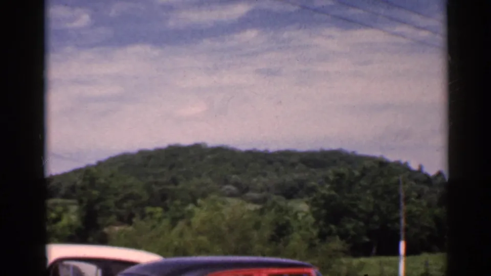 1959 Indiana 8mm frame with a red car crossing the foreground beneath wooded hills, field, sky, and a roadside utility pole.