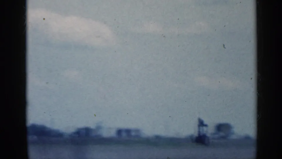 Soft 1959 Indiana 8mm view of blue sky over a distant rural horizon, with indistinct buildings or structures near the field edge.