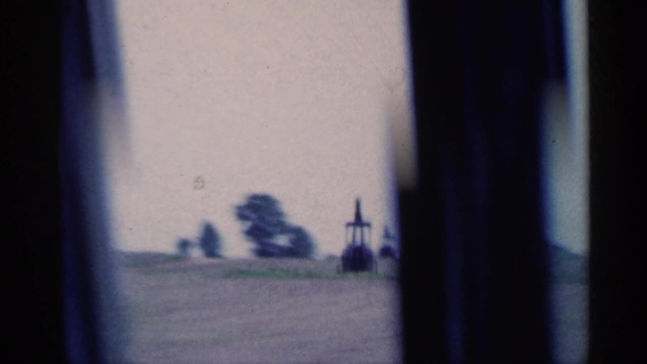1959 Indiana farmland passes by with a distant oil pumpjack, tree line, hazy sky, and dark window edges from the moving car.