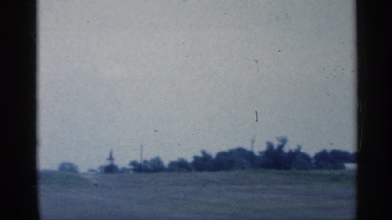 A second moving-car view of Indiana fields shows a distant pumpjack near a low tree line under a hazy 1950s sky.