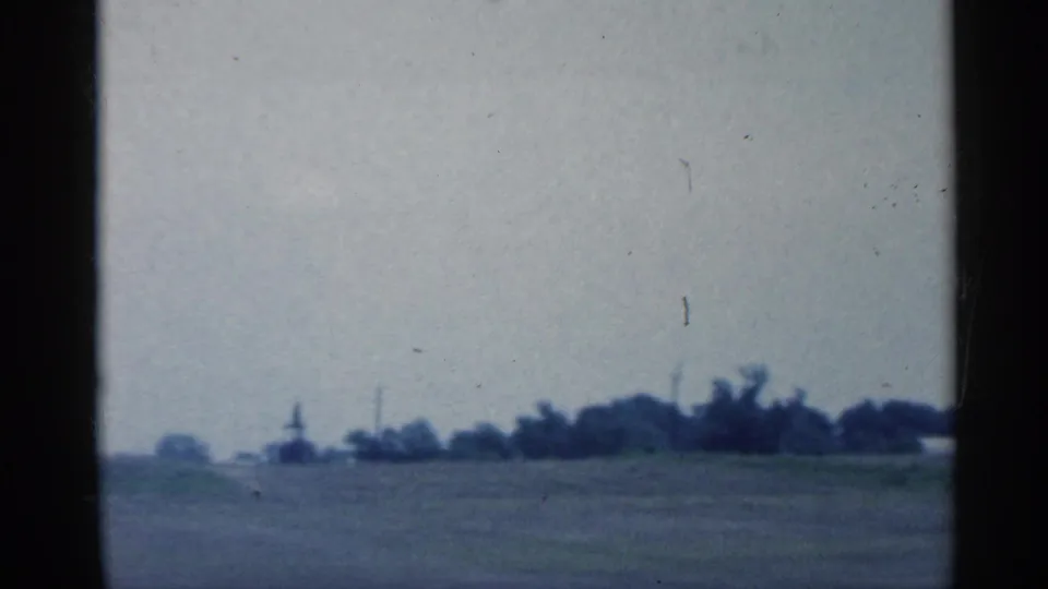 A second moving-car view of Indiana fields shows a distant pumpjack near a low tree line under a hazy 1950s sky.