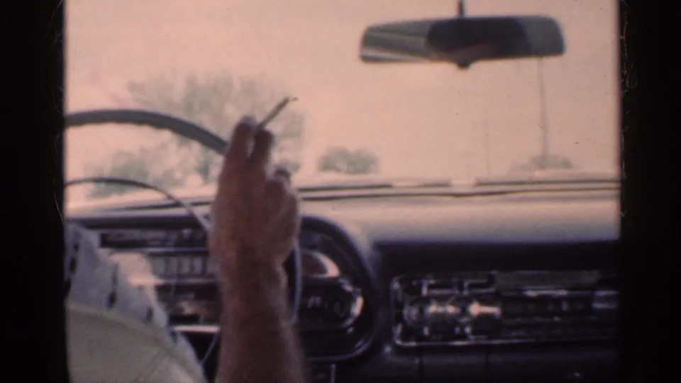 A hand holds a cigarette inside a classic car, with the steering wheel, dashboard controls, rearview mirror, and hazy rural view visible.