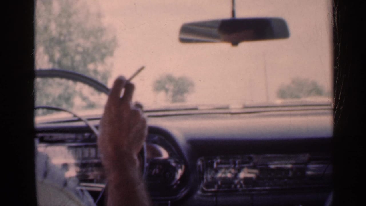 Driver's hand holding a cigarette beside the steering wheel inside a moving car.