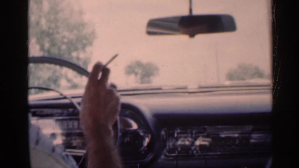 1959 Indiana home-movie frame from inside a car, with a driver's hand holding a cigarette near the steering wheel and dashboard.