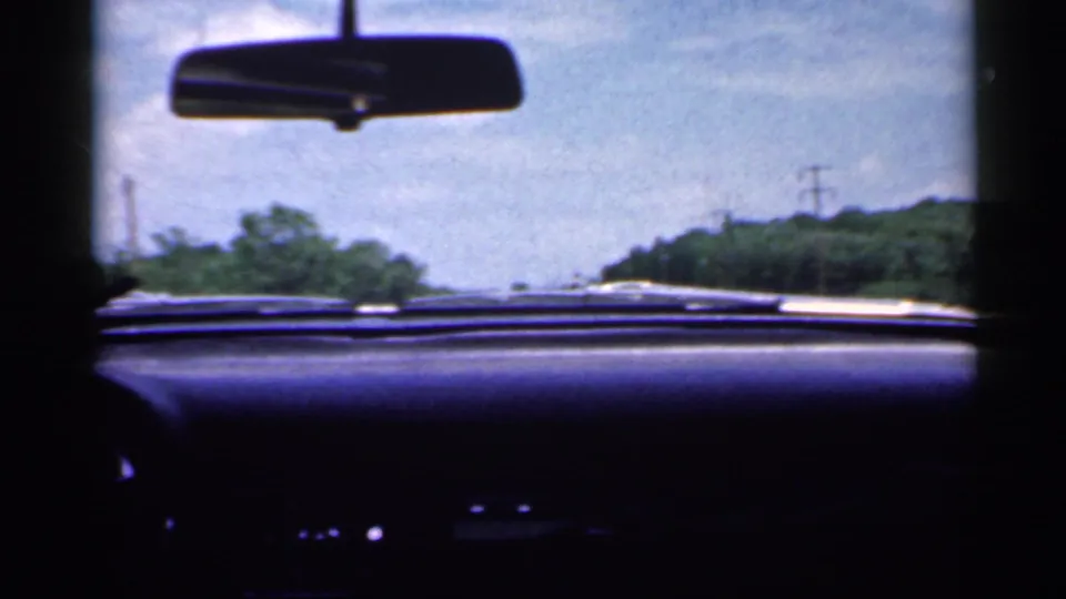 1959 Indiana windshield view over the dashboard toward a rural road, roadside trees, blue sky, and a rearview mirror.