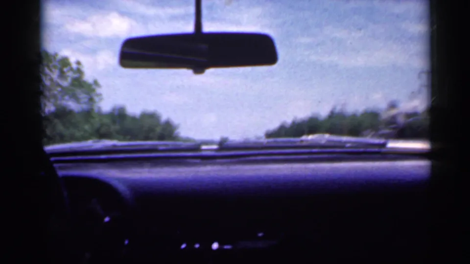 Late-1950s Indiana road-trip frame looking past the dashboard and rearview mirror toward a tree-lined rural road.