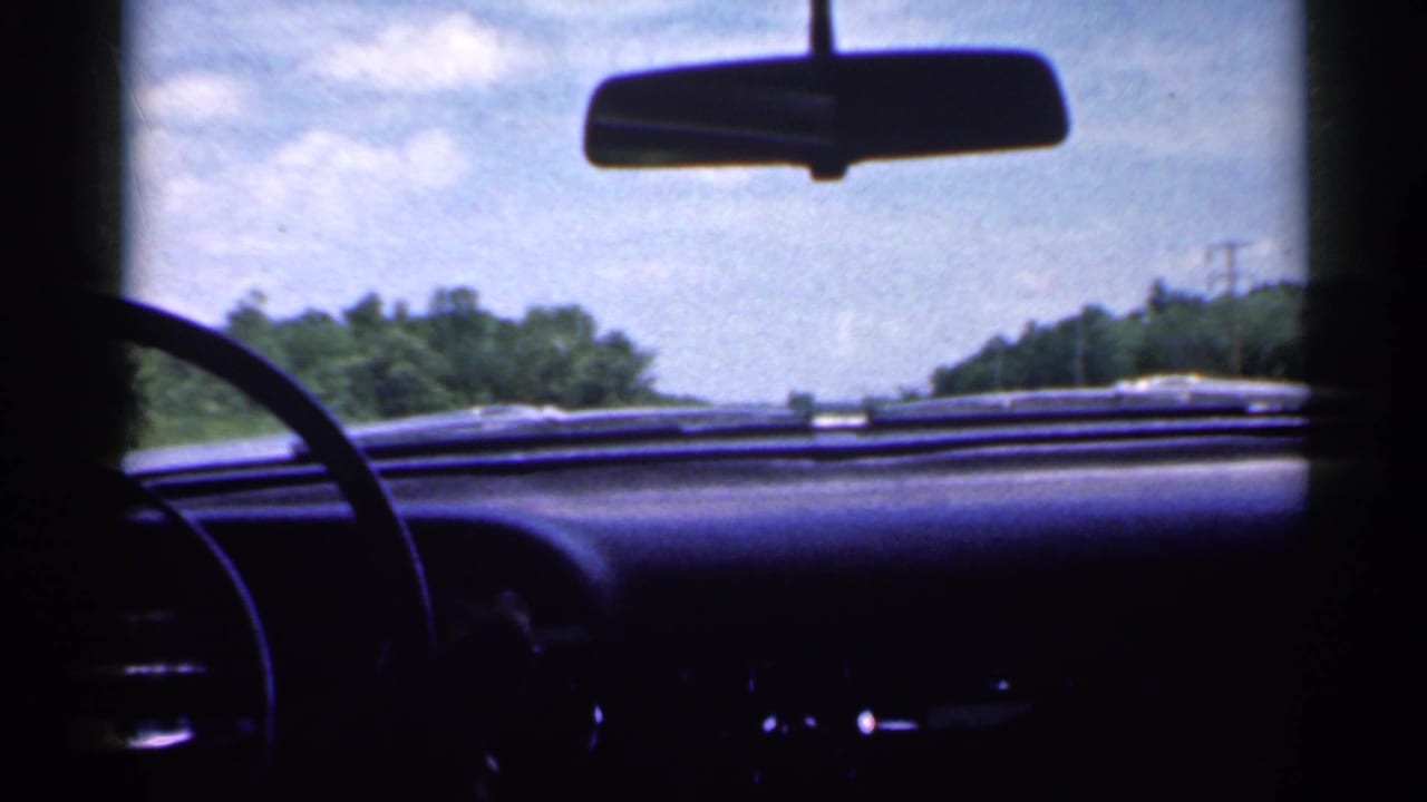 Car interior facing a rural road through the windshield with dashboard, steering wheel, and rearview mirror visible.