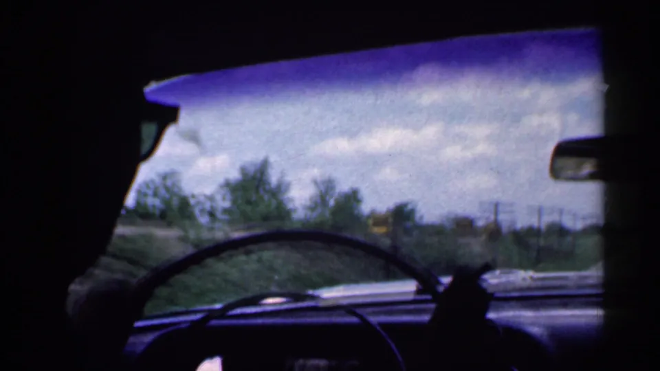 1959 Indiana 8mm frame from inside the car, showing the driver's silhouette, steering wheel, windshield, roadside trees, sky, and power lines.