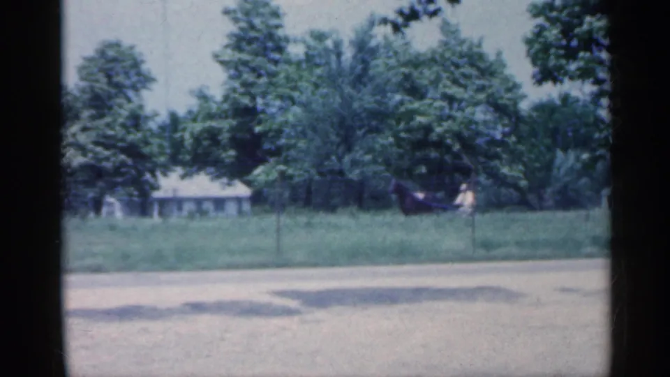 The horse-drawn carriage continues across a rural Indiana field, with a road edge in front and houses partly visible through trees.