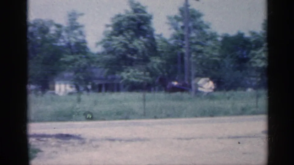 A distant driver in a light shirt guides a horse-drawn carriage through tall grass near trees and a small Indiana rural house.
