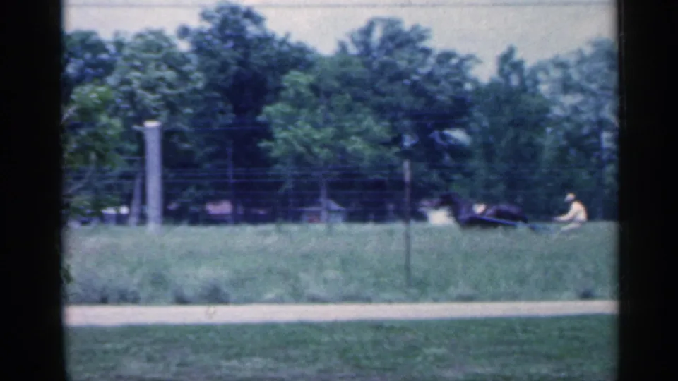 The horse-drawn cart appears farther right in the rural Indiana frame, passing near a fence and tall grass.