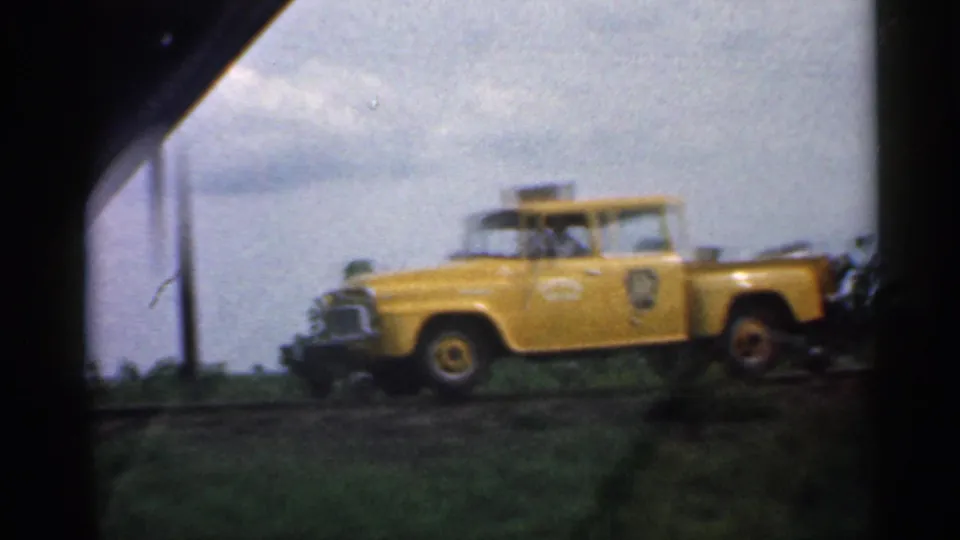 A yellow vintage pickup, possibly a service vehicle, is seen beside a railroad crossing in the 1959 Indiana home movie.