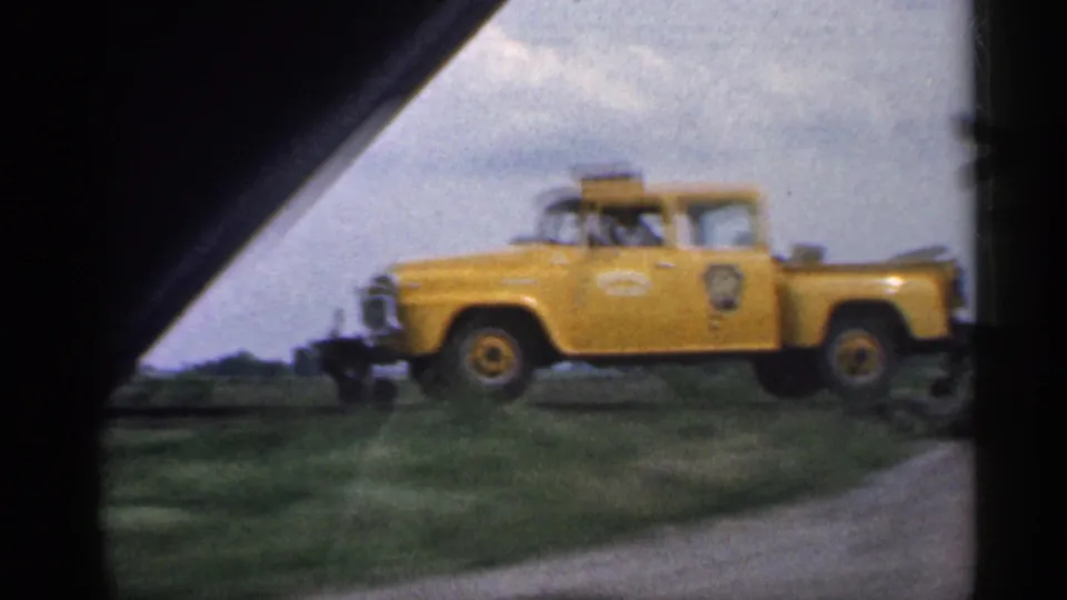 1959 Indiana 8mm frame showing a yellow utility truck with door markings moving past grass fields, filmed from inside a car.