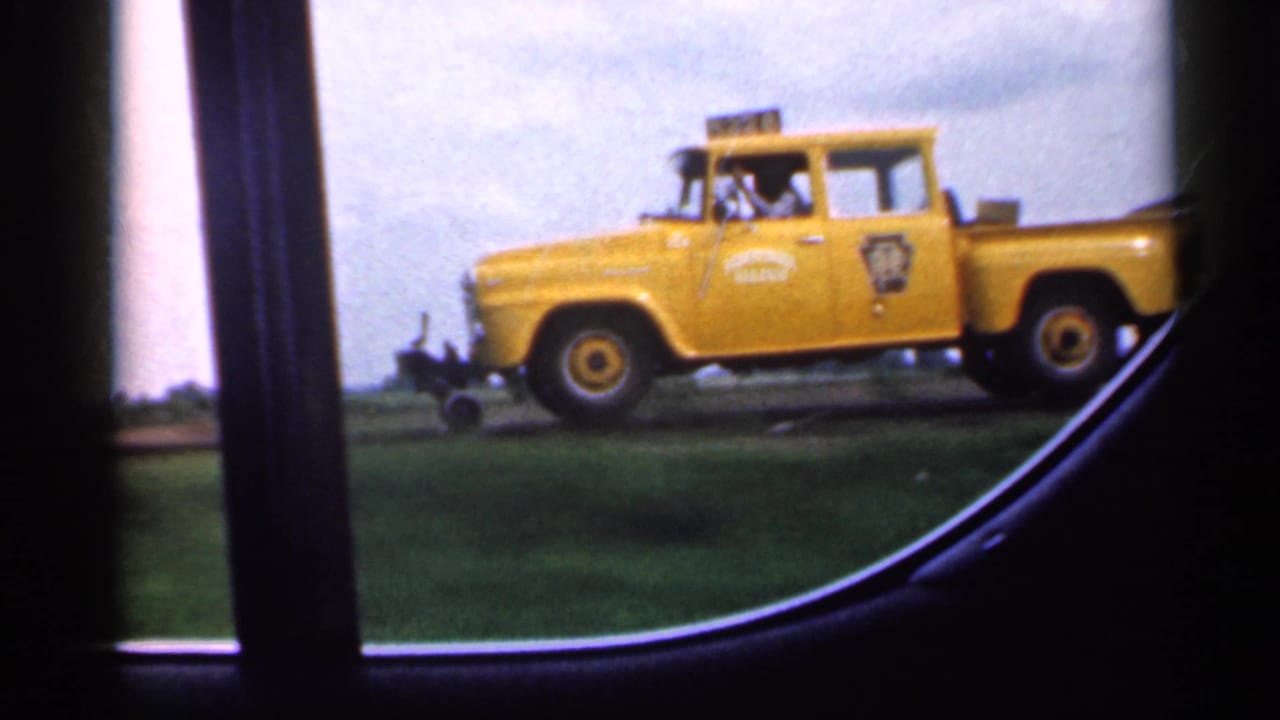 Yellow marked truck framed by a car window with fields behind it.