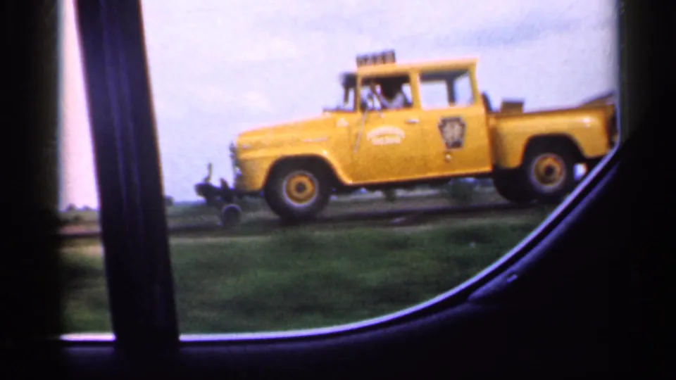 1959 Indiana 8mm still with the yellow utility truck centered behind a dark car-window pillar, with fields and sky in the background.