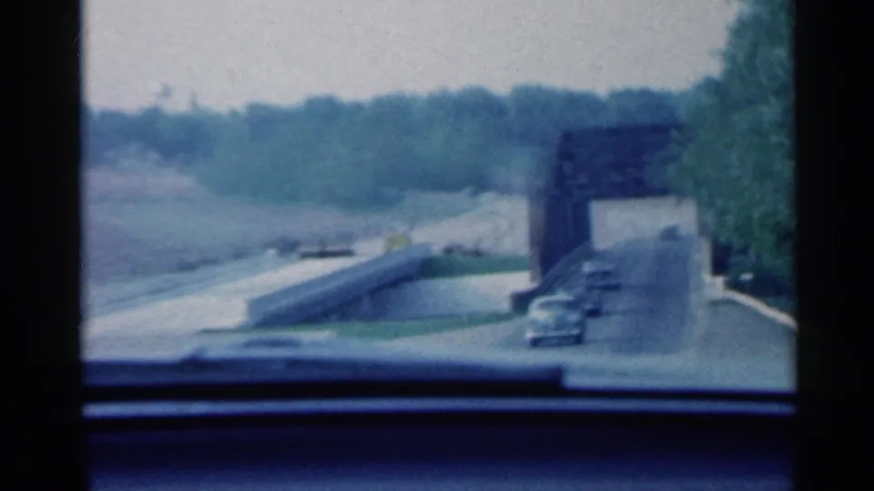 1959 Indiana road scene from inside a moving car, approaching a metal truss bridge with another car ahead.