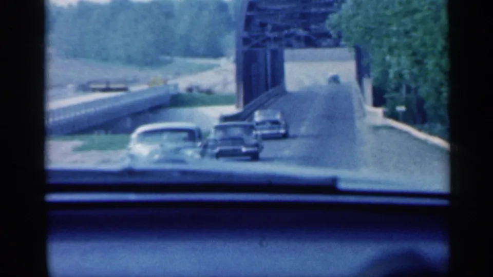 1959 Indiana 8mm frame showing multiple mid-century cars near a metal truss bridge, filmed from inside a moving vehicle.