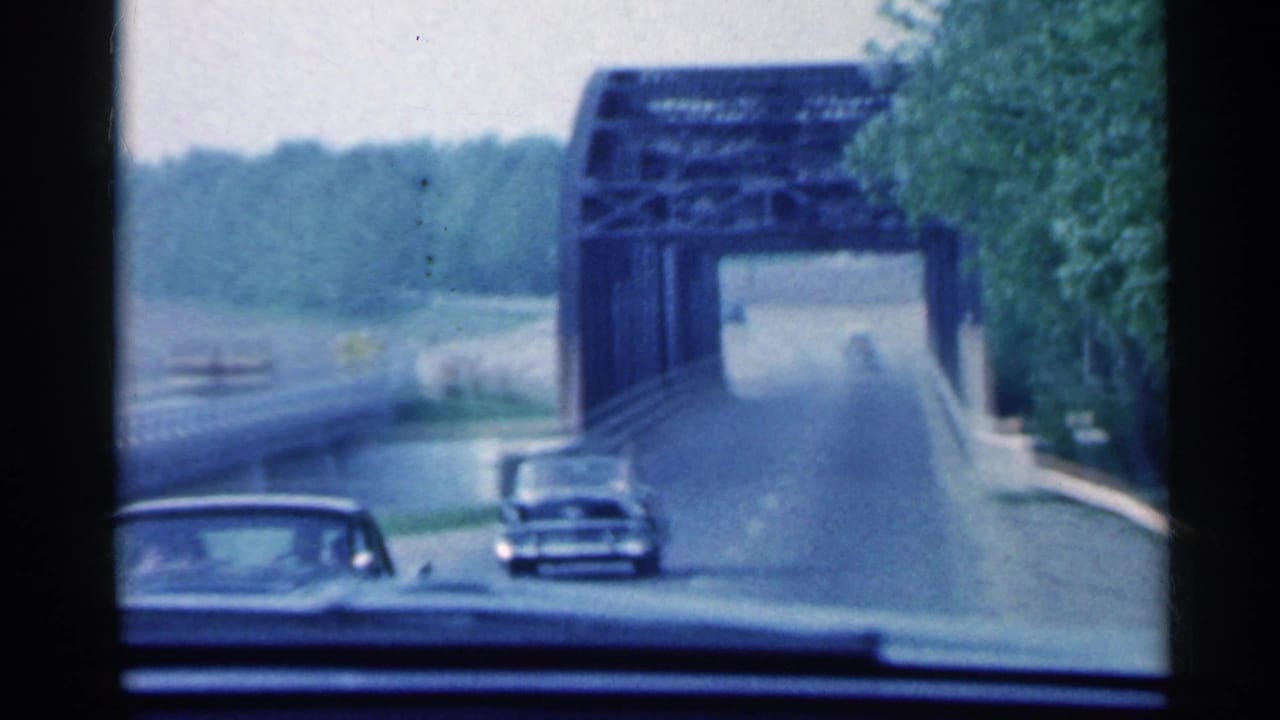 Windshield view of cars approaching a steel truss bridge on a tree-lined road.