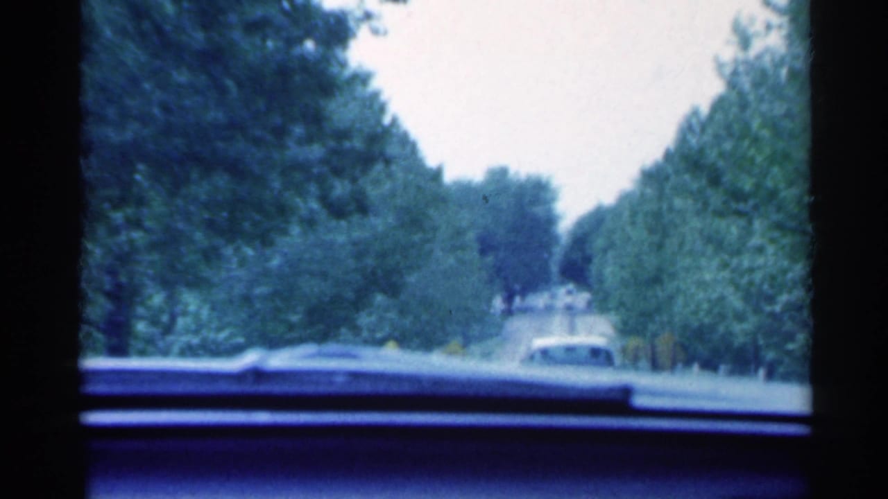 A moving-car view in 1959 Indiana shows a vehicle ahead on a tree-lined roadway through the windshield.