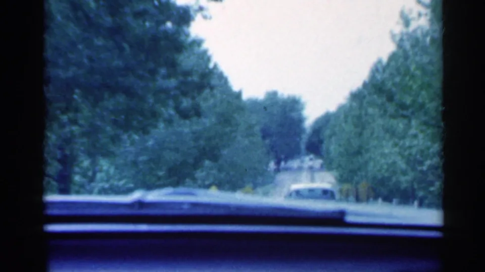A moving-car view in 1959 Indiana shows a vehicle ahead on a tree-lined roadway through the windshield.