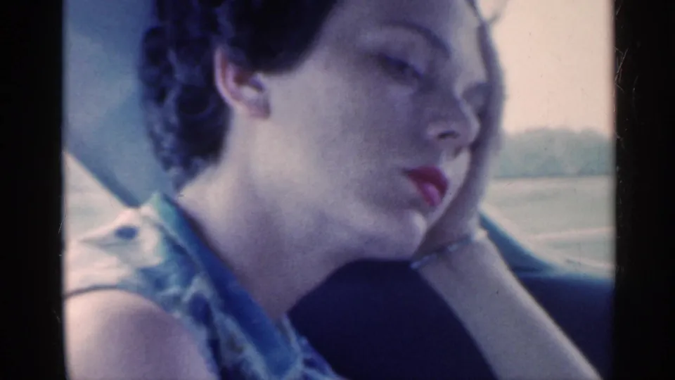 In the 1959 home movie, a woman inside the car rests her head on her hand while looking tired or contemplative.