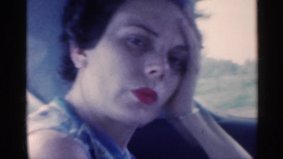 In this 1959 Indiana 8mm frame, a woman sits in a car and looks toward the camera while resting her head on her hand.
