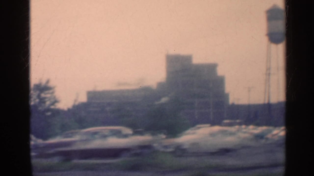 1959 Indiana 8mm frame showing a water tower, passing cars, and distant downtown buildings through soft motion blur.