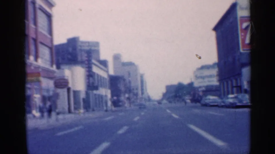 1959 Indiana 8mm frame looking down a downtown street with storefronts, signs, parked cars, and a hazy distance.