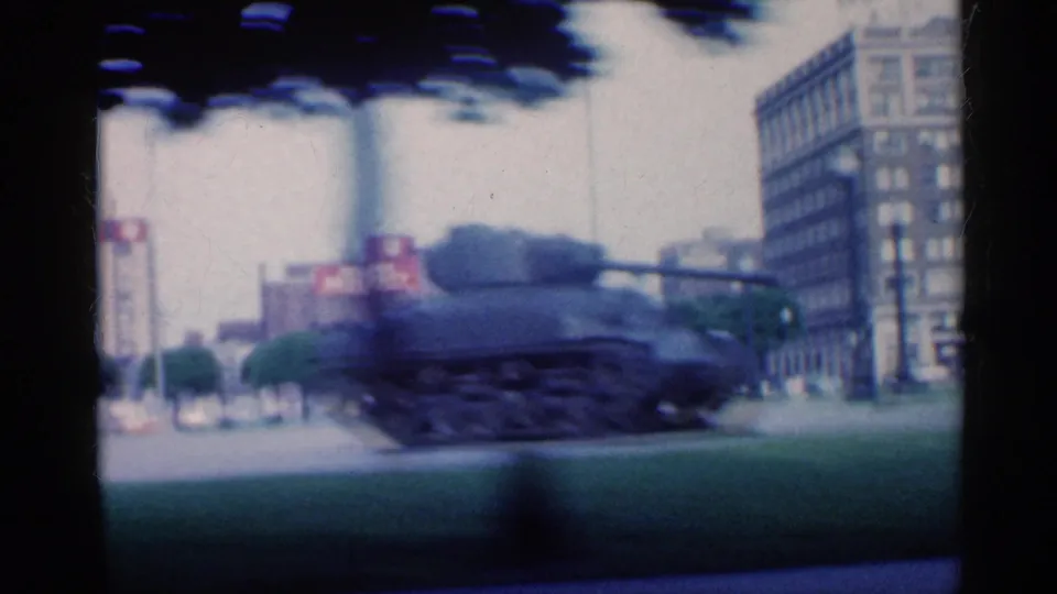 1959 Indiana home-movie frame showing a tank-like vehicle passing a park area, with city buildings and trees in the background.
