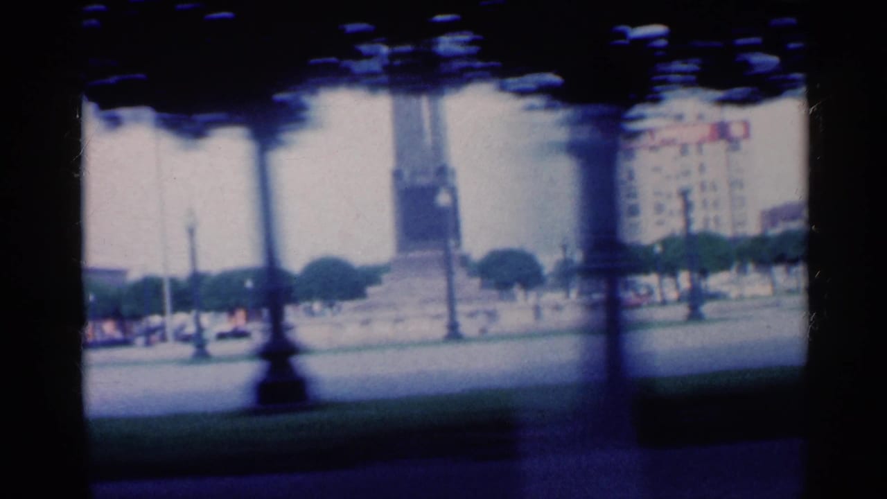 Moving view past a city square with a tall monument, lawn, trees, and distant buildings.