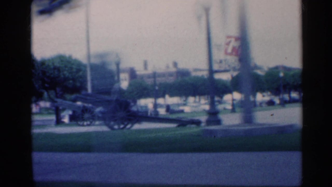 Display cannon on a lawn near a monument, trees, and a distant commercial sign.
