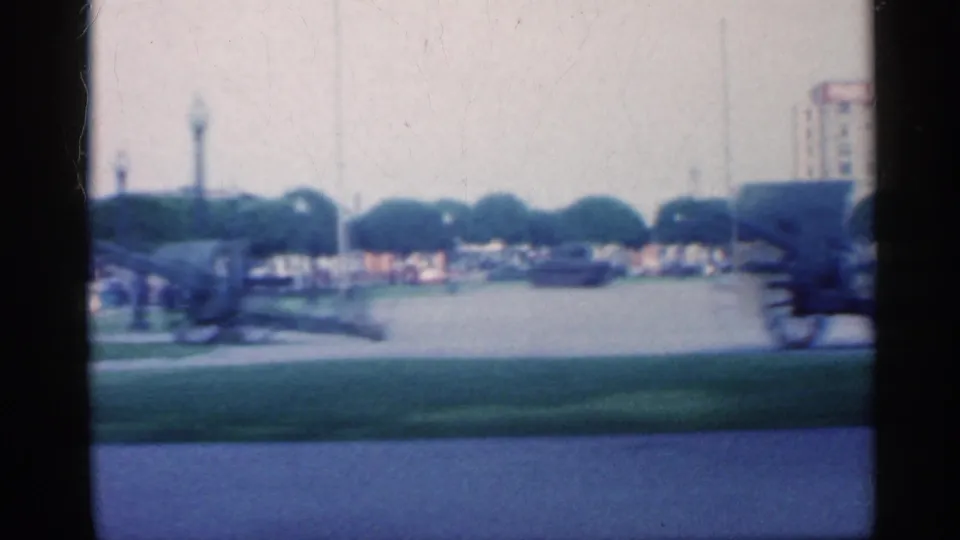 Vehicle-shot 1959 Indiana streetscape with a cannon display, open lawn, trimmed trees, and downtown buildings in soft focus.