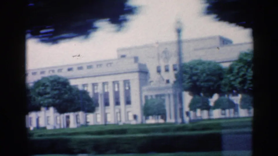 1959 Indiana home-movie frame showing a large civic-looking building, monument column, trees, lawn, and street foreground.