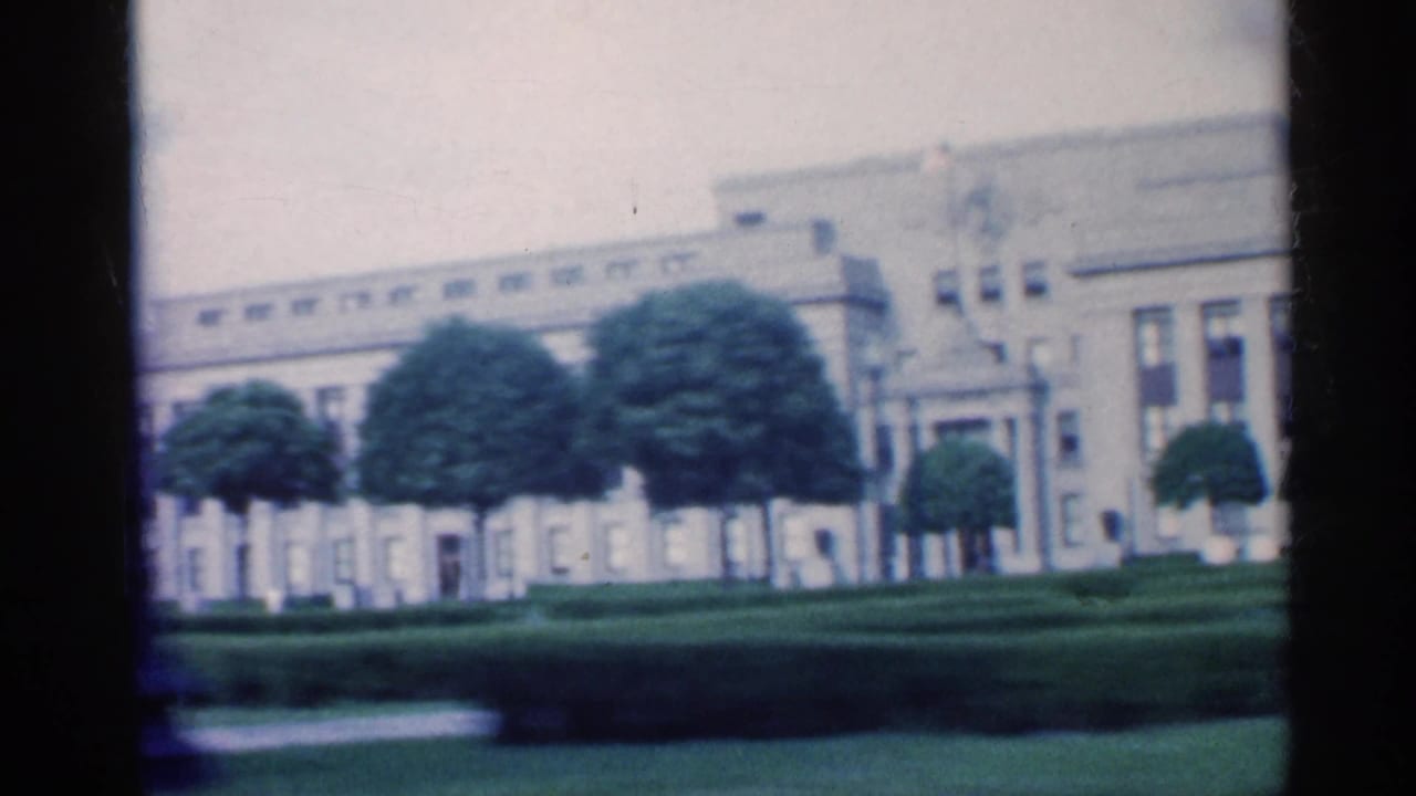 1959 Indiana 8mm frame showing a large formal building across landscaped grass, hedges, and trees.