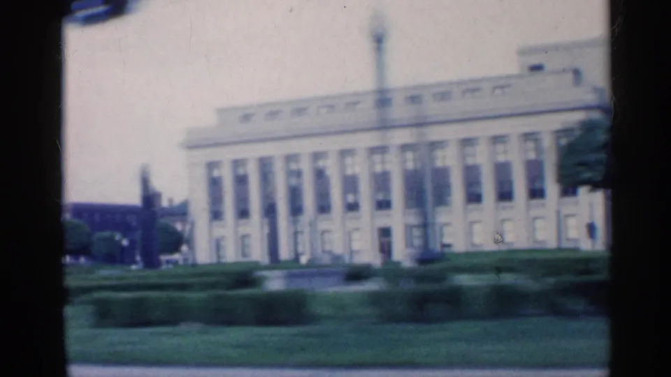 1959 Indiana home-movie frame of a columned institutional-looking facade beyond clipped hedges and grass.