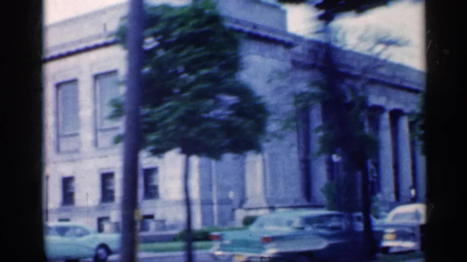 Street-level 1959 Indiana frame with parked mid-century cars beside a large stone building and trees.