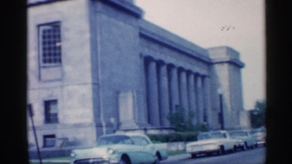 1959 Indiana 8mm frame showing the side of a large columned building with mid-century cars parked at the curb.