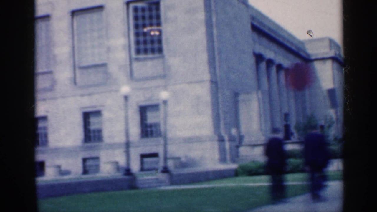 1959 Indiana 8mm frame showing a pale classical building exterior, lawn edge, windows, and two dark-suited pedestrians passing in soft focus.