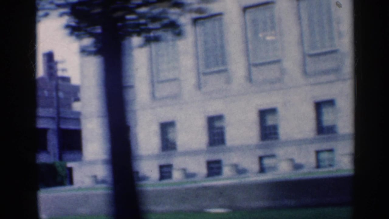 1959 Indiana home-movie frame with a tree trunk crossing a pale institutional-looking building facade, windows, lawn, and sidewalk.