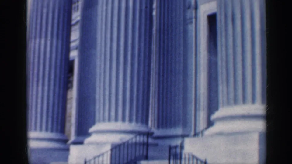 1959 Indiana 8mm frame showing classical columns, a pale facade, railings near an entrance, and part of a vintage car at the lower right.