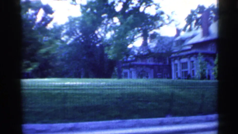 1959 Indiana 8mm frame of a residential house partly screened by trees beyond a broad lawn and road edge.